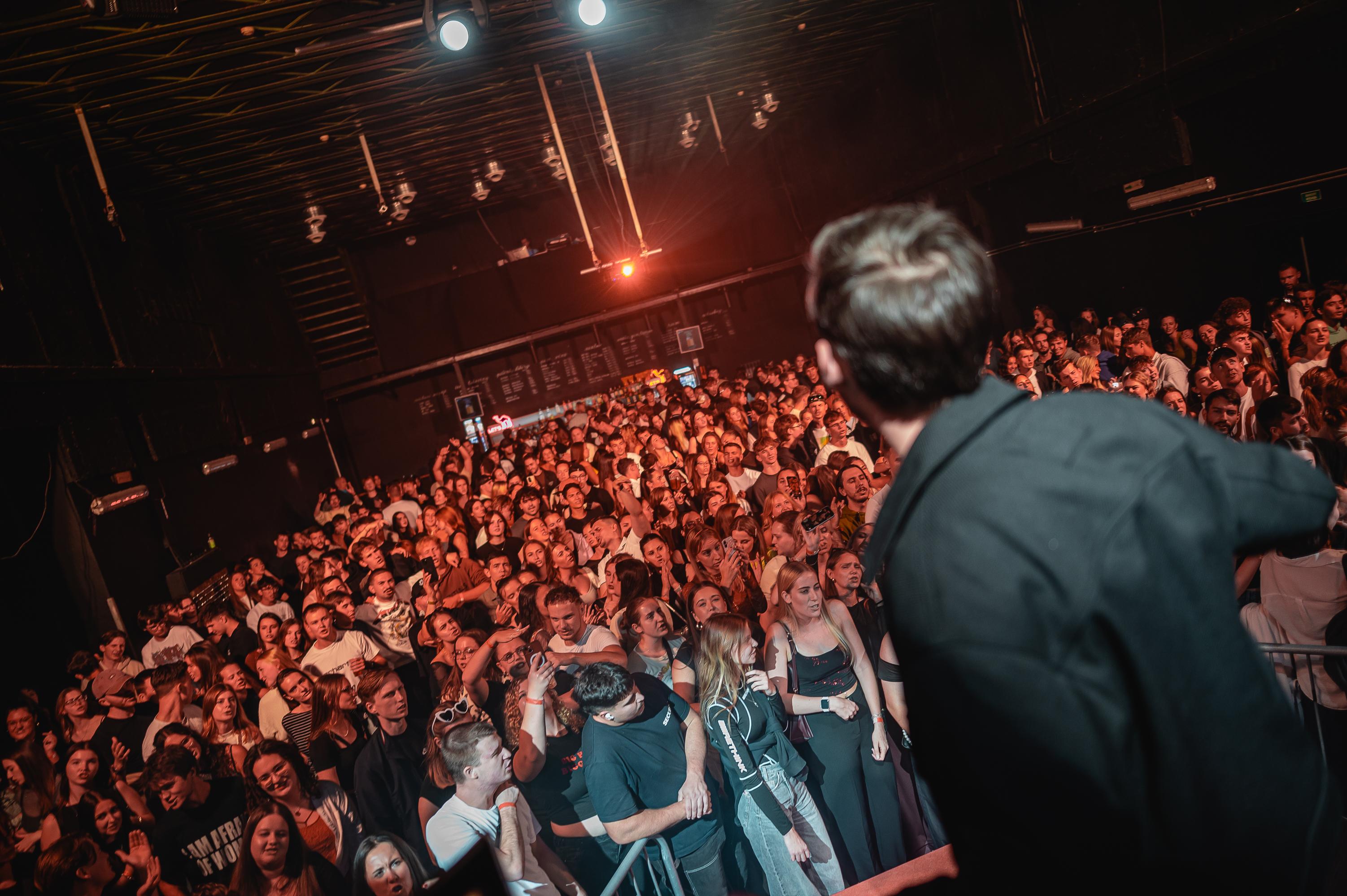 Zpěvák zezadu na klubovém pódiu Karaoke Party v Brně, před ním plný sál v teplém klubovém světle