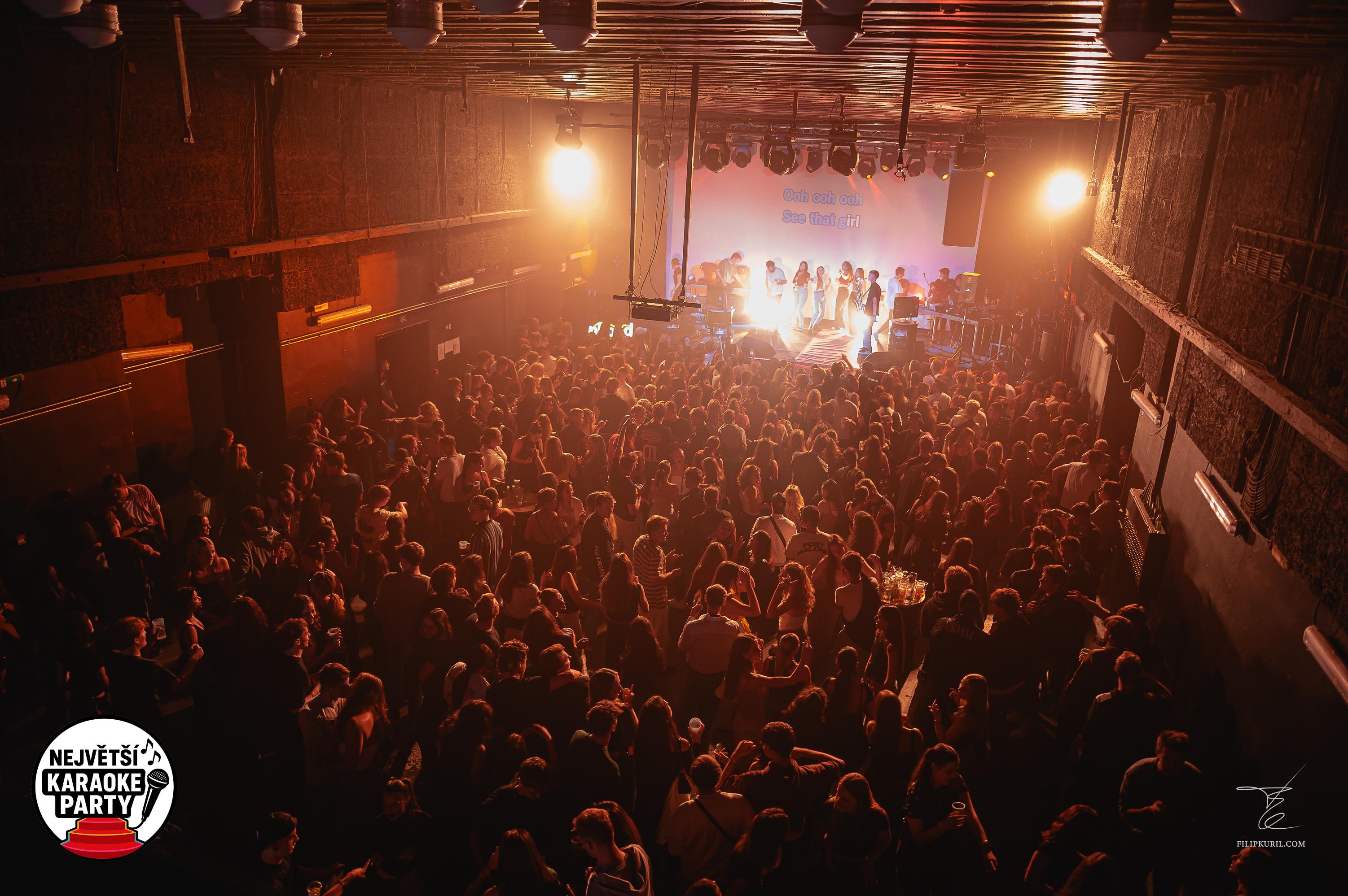 Vyprodaný klub Karaoke Party s davem stovek lidí, jeviště zalité teplým světlem, panel s logem „Největší karaoke party v Česku" v rohu