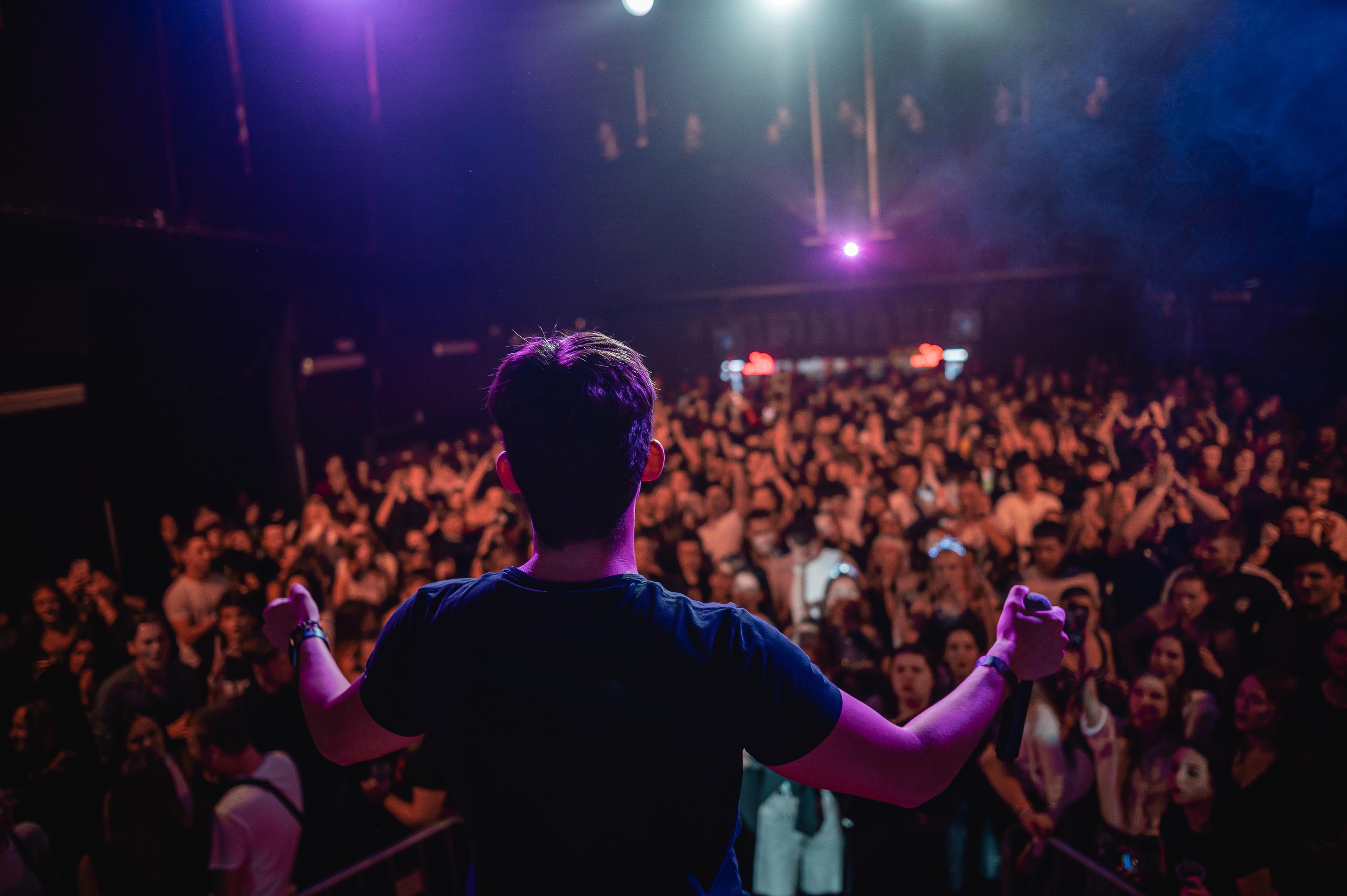 Vyprodaný klub Karaoke Party v Praze — zpěvák zezadu s rozpaženýma rukama nad davem pod fialovými reflektory
